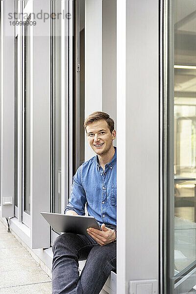 Junger Geschäftsmann  der ein digitales Tablet benutzt  während er an der Bürotür sitzt