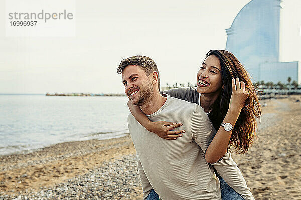 Freund lächelt  während er seine Freundin am Strand huckepack nimmt
