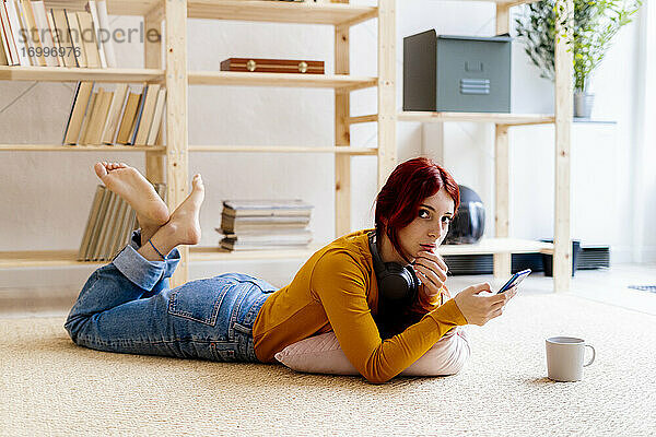 Rothaarige Frau  die ein Mobiltelefon benutzt  während sie zu Hause auf dem Teppich liegt