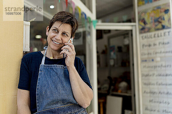 Älterer Ladenbesitzer  der an der Tür einer Werkstatt steht und mit einem Mobiltelefon spricht