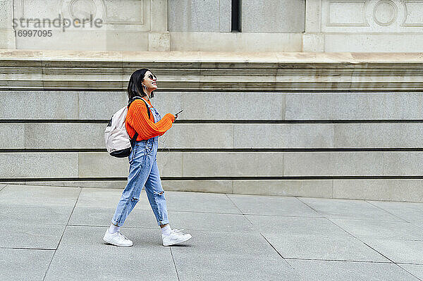 Junge Frau mit Rucksack und Mobiltelefon  die beim Gehen auf dem Fußweg wegschaut