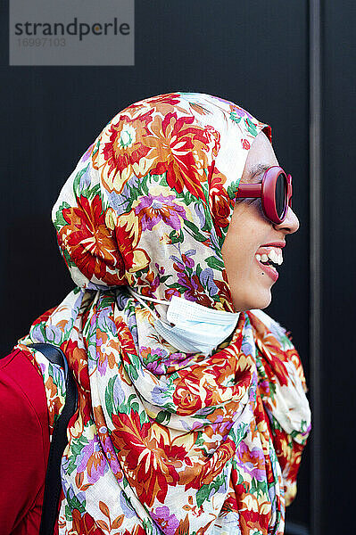 Fröhliche Frau mit geblümtem Hidschab vor schwarzer Wand während der COVID-19