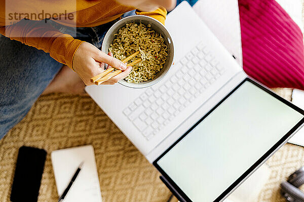 Junge Frau isst Nudeln und studiert zu Hause am Laptop
