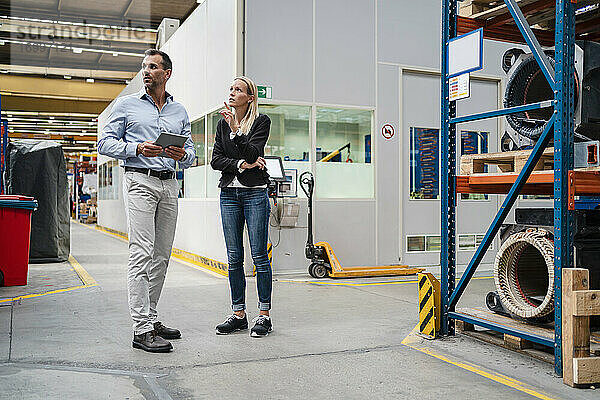 Ein Geschäftsmann hält ein digitales Tablet  während eine Unternehmerin in einer Fabrik daneben steht