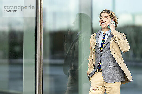 Glücklicher Geschäftsmann mit Hand in der Tasche  der mit einem Mobiltelefon spricht  während er an einer Glaswand steht