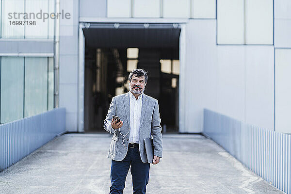 Lächelnder Geschäftsmann mit Smartphone und Kopfhörern vor einem Bürogebäude