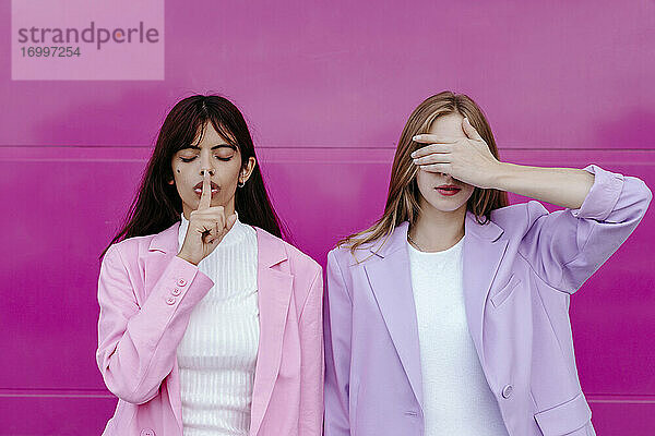 Junge Frau mit Finger auf den Lippen  die neben ihrer Schwester steht und die Augen vor einer rosa Wand verdeckt