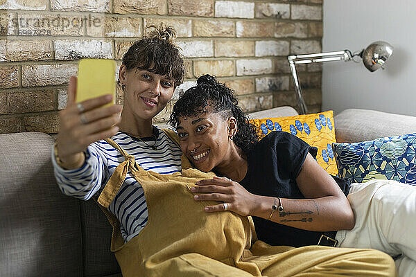 Schwangere Frau macht Selfie auf dem Handy mit ihrer Freundin  während sie zu Hause auf dem Sofa sitzt