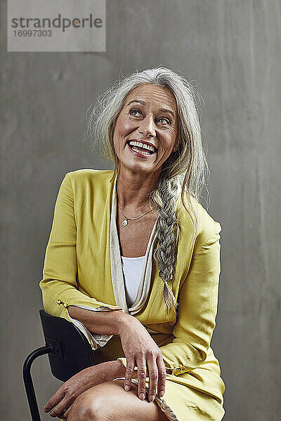 Glückliche Unternehmerin blickt auf  während sie auf einem Stuhl vor einer grauen Wand sitzt