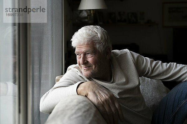 Lächelnder älterer Mann  der durch das Fenster schaut  während er zu Hause auf dem Sofa sitzt