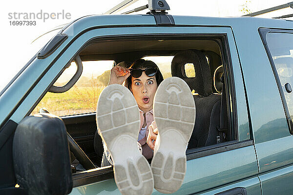 Überrascht junge Frau sitzt mit Füßen oben auf Autofenster während Sonnenuntergang