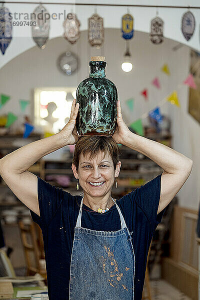 Lächelnde Künstlerin mit türkisfarbener Flasche auf dem Kopf im Keramikladen