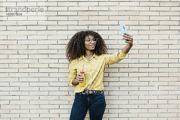 Junge Frau lächelnd bei der Aufnahme von Selfie durch Handy stehen gegen Backsteinmauer