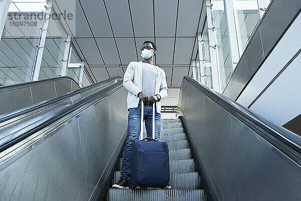 Geschäftsmann mit Gesichtsschutzmaske  der ein Gepäckstück hält  während er auf einer Rolltreppe im Bahnhof steht