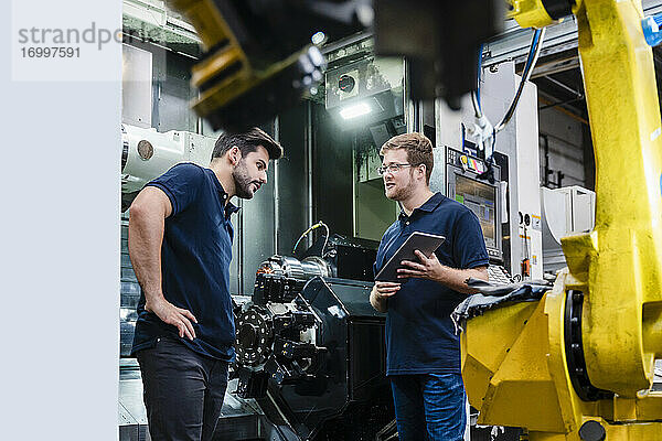 Männliche Mitarbeiter diskutieren über einen Roboterarm  während sie ein digitales Tablet in einer Produktionsstätte halten