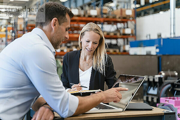 Eine lächelnde Kollegin und ein Geschäftsmann benutzen einen Laptop  während sie in einer Fabrik stehen