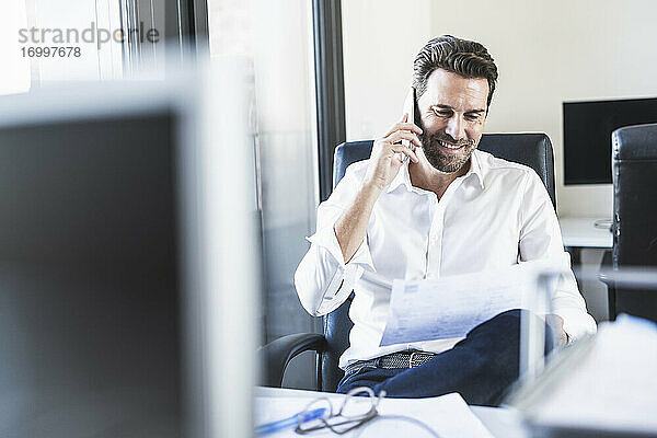 Geschäftsmann  der auf Papier schaut  während er im Büro sitzt und mit seinem Handy spricht