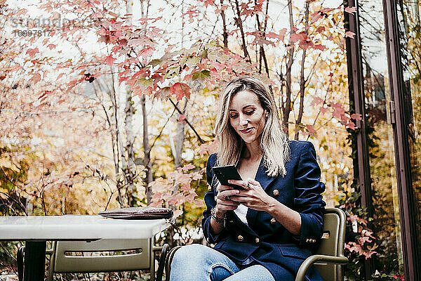 Blonde Frau im mittleren Erwachsenenalter  die ein Smartphone benutzt  während sie auf der Terrasse vor den Herbstbäumen sitzt
