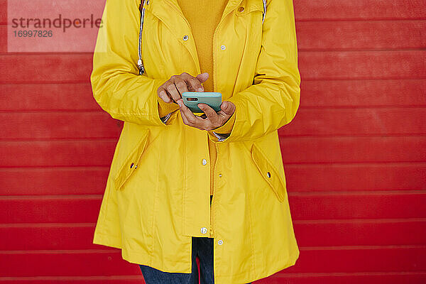 Frau in gelbem Regenmantel mit Smartphone vor roter Wand