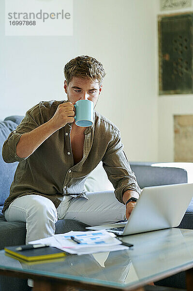 Geschäftsmann trinkt Kaffee  während er zu Hause am Laptop arbeitet