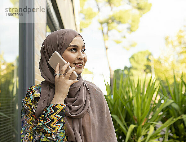 Junge Frau mit Hidschab  die im Freien steht und mit einem Mobiltelefon spricht
