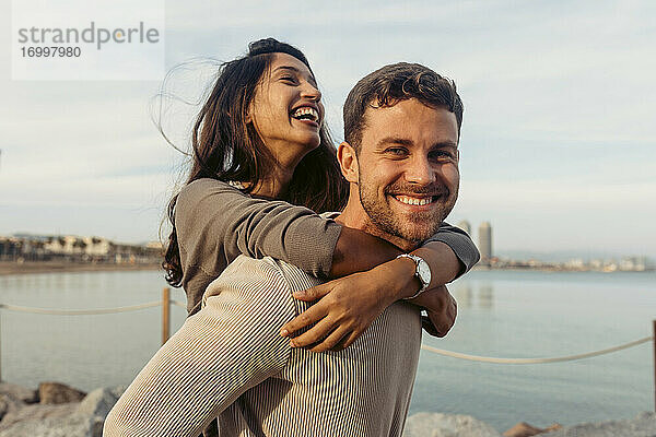 Junger Mann lächelt  während er eine lachende Frau am Strand huckepack nimmt