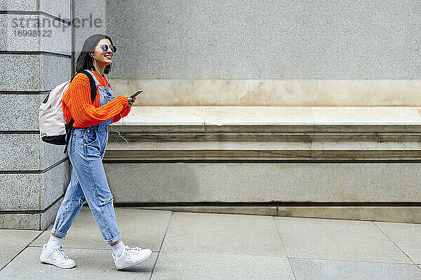 Lächelnde Frau mit Rucksack  die ein Mobiltelefon benutzt  während sie gegen eine Wand läuft