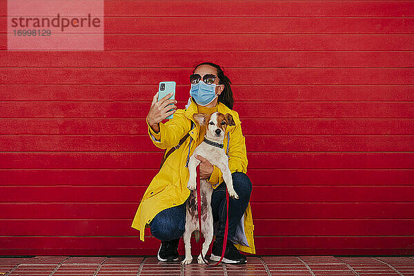 Frau mit Gesichtsmaske und Hund  die Smartphone-Selfies vor einer roten Wand macht