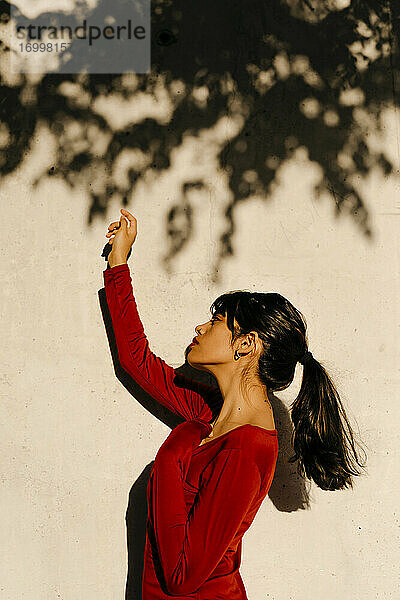 Schöne Frau mit erhobener Hand  stehend vor dem Schatten eines Baumes an der Wand