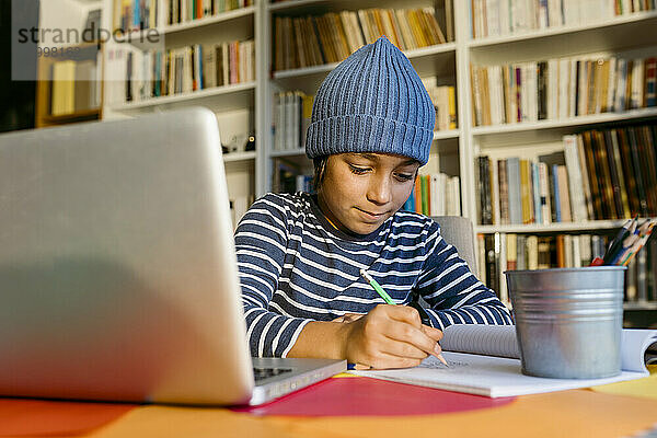 Lächelnder Junge mit Strickmütze  der in ein Buch schreibt  während er zu Hause sitzt
