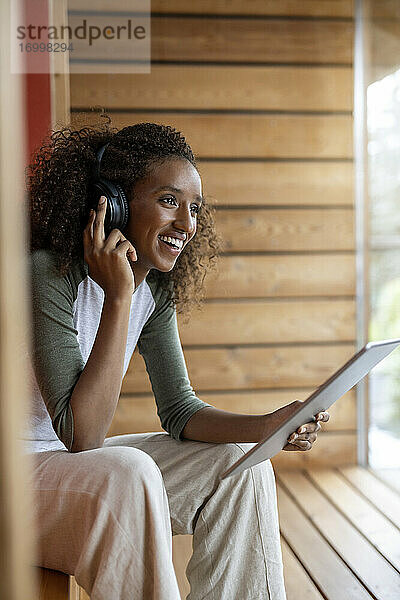Lächelnde Frau  die über Kopfhörer spricht  während sie zu Hause ein digitales Tablet benutzt