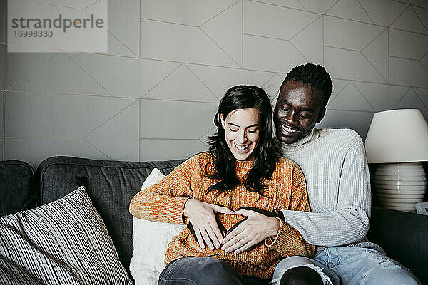 Glücklicher Mann und schwangere Frau bilden Herzform mit Händen auf dem Bauch  während sie auf dem Sofa zu Hause sitzen