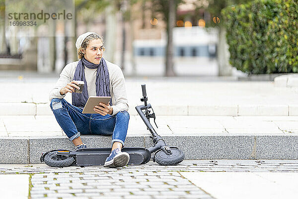 Mann mit Elektroroller  der auf dem Gehweg sitzend ein digitales Tablet benutzt