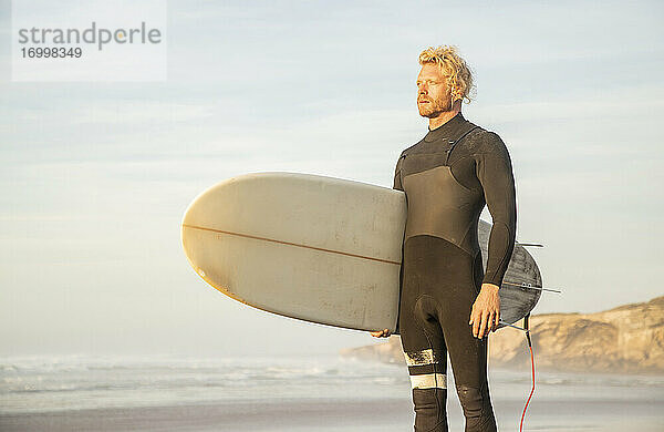Blonder Mann im Neoprenanzug  der ein Surfbrett trägt  während er am Strand gegen den Himmel schaut
