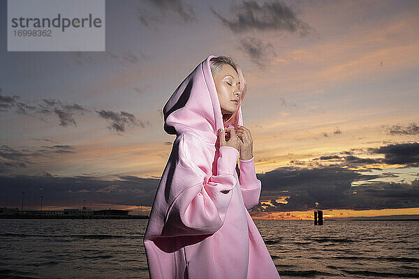 Junge Frau mit rosa Kapuzenshirt steht bei Sonnenuntergang am Strand