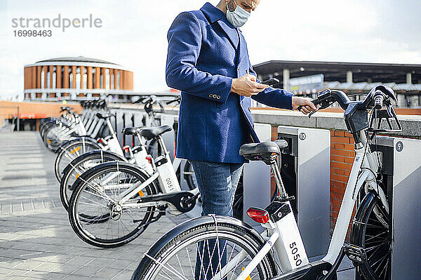 Älterer Mann mit Smartphone an einer Fahrradparkstation während COVID-19