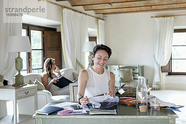 Lächelnde Geschäftsfrau  die einen Stoff auswählt  während eine Kollegin im Büro zu Hause mit ihrem Smartphone telefoniert