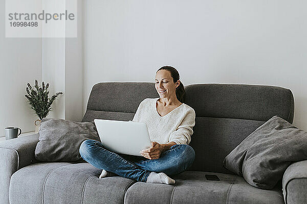 Lächelnde Frau  die vom Laptop aus einen Videogespräch führt  während sie zu Hause auf dem Sofa sitzt