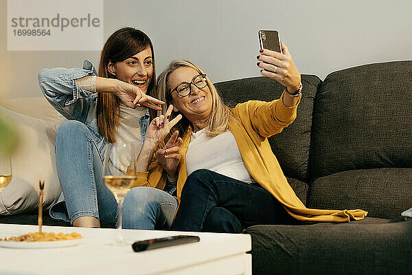 Lächelnde Mutter und Tochter  die ein Friedenszeichen machen  während sie ein Selfie mit dem Smartphone zu Hause machen