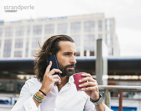 Unverfälschtes Porträt eines bärtigen Geschäftsmannes  der im Freien sitzt und mit seinem Smartphone spricht