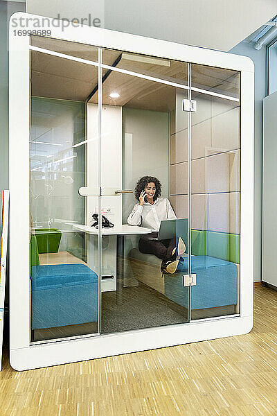 Geschäftsfrau  die mit einem Mobiltelefon spricht  während sie an einem Laptop in einer Kabine im Büro arbeitet