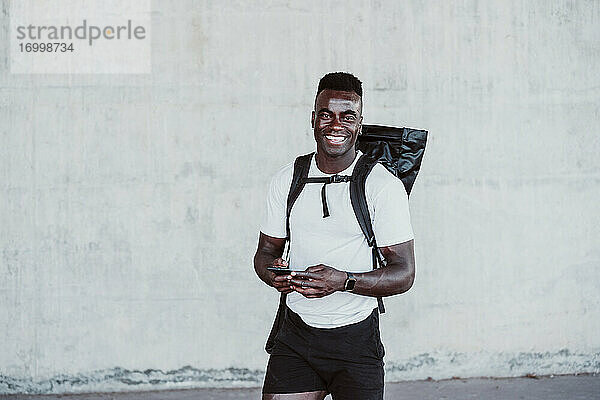 Lächelnder männlicher Sportler mit Smartphone an der Wand stehend