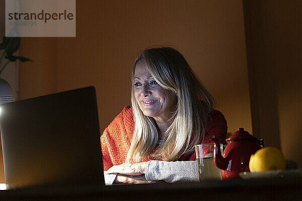 Frau lächelt  während sie einen Videoanruf auf dem Laptop hört  während sie zu Hause sitzt