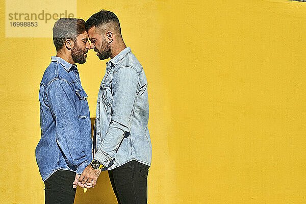 Schwule Männer halten sich an den Händen  während sie von Angesicht zu Angesicht an einer gelben Wand stehen