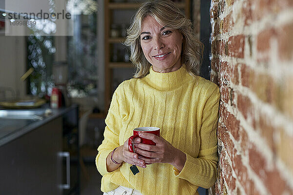 Lächelnde reife Frau lehnt an einer Backsteinmauer und hält eine Teetasse in der Küche