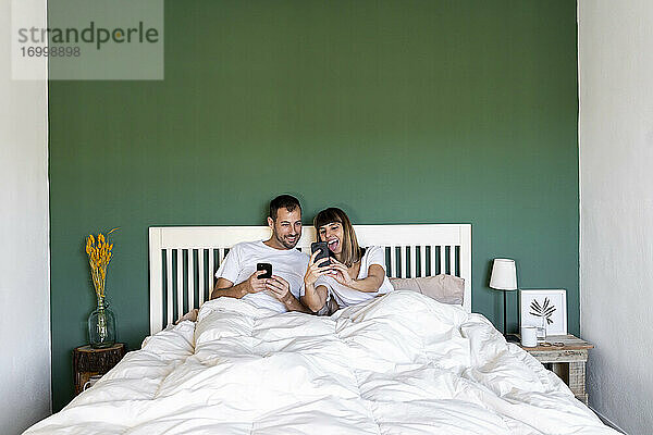 Glückliche Frau  die einem Mann  der zu Hause auf dem Bett sitzt  ihr Mobiltelefon zeigt