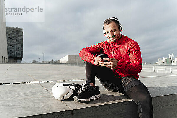 Lächelnder junger Mann  der im Freien sitzend Musik hört und ein Mobiltelefon benutzt