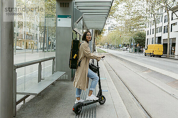 Frau mit Tretroller und Instrumentenkoffer an einer Straßenbahnhaltestelle in der Stadt