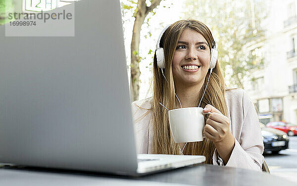 Frau mit Kopfhörern trinkt Kaffee  während sie mit einem Laptop in einem Straßencafé sitzt