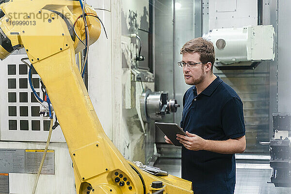 Männlicher Arbeiter  der einen Roboterarm untersucht  während er ein digitales Tablet in einer Fabrik hält